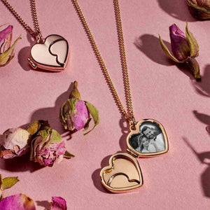 A 14K solid gold heart-shaped photo locket medallion from the Timeless Collection hangs gracefully from a delicate chain. The handcrafted locket features a smooth, reflective surface with a curved line design that suggests it can open. Hypoallergenic and elegantly styled, this exquisite piece is beautifully displayed against a white background.