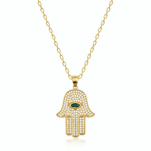 The 14K Solid Gold Emerald Hamsa Necklace showcases a pendant with a central eye-shaped green stone, providing protection against the evil eye. The design is beautifully intricate, highlighted by small clear stones, and complemented by a simple yet elegant chain.