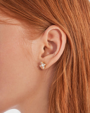 The 14K Solid Gold Theia Diamond Natural Pearl Dot Stud Earrings from the Elegance Collection feature a central pearl encircled by four gold prongs, each embellished with small lab-grown diamonds, creating a cross-like design. Crafted from hypoallergenic 14K real solid gold, one earring is displayed from the front and the other from the back.