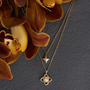 Part of the Elegance Collection, the 14K Solid Gold Natural Pearl Double Layer Necklace features a hypoallergenic design with two layers: the upper chain displays a small diamond-shaped pendant while the lower chain highlights an intricate intertwined design holding a natural pearl, all set against a white background.