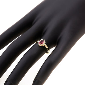 The 14K Solid Gold Ruby July Birthstone Ring features an oval red gemstone encircled by a halo of small, round diamonds. The band is sleek, polished, and stamped with the 14K mark on the inside. Each ring is part of our exclusive handmade jewelry collection and is presented against a plain white background.