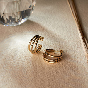 A pair of 14K Solid Real Gold Triple Ropes Hoop Earrings is showcased, with one earring standing upright while the other rests flat. This composition highlights its round shape and intricate clasp detail against a pristine white background—perfectly capturing elegance and simplicity.