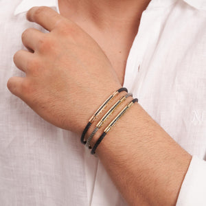 This elegant men's accessory, known as the 14K Solid Gold Men's Greek Key Bracelet, features a stylish twisted black cable band with a 14K solid gold clasp. It is highlighted by a gold bar adorned with small, dark stones arranged in a sophisticated linear pattern.