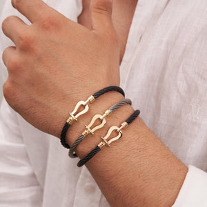 A braided rope bracelet in sleek black featuring a 14K solid gold clasp styled like a shackle. This design marries elegance with simplicity, making it suitable for both casual and formal occasions and an ideal choice for fans of sophisticated handcuff jewelry.