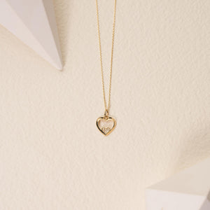 The Real Diamond Heart Necklace in 14K Gold features a delicate design with a heart-shaped pendant. Inside this stunning pendant, a smaller heart sparkles with natural diamonds. The elegant gold chain elegantly drapes against the white backdrop, exuding sophistication and allure.