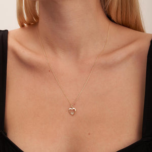 The Real Diamond Heart Necklace in 14K Gold features a delicate design with a heart-shaped pendant. Inside this stunning pendant, a smaller heart sparkles with natural diamonds. The elegant gold chain elegantly drapes against the white backdrop, exuding sophistication and allure.