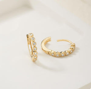 The Genuine Diamond Hoop Earrings in 14K Solid Gold include one earring lying flat and the other standing upright. These elegant earrings are crafted with genuine gold and adorned with a row of small, round white diamonds along the outer edge, giving them a shimmering appearance.