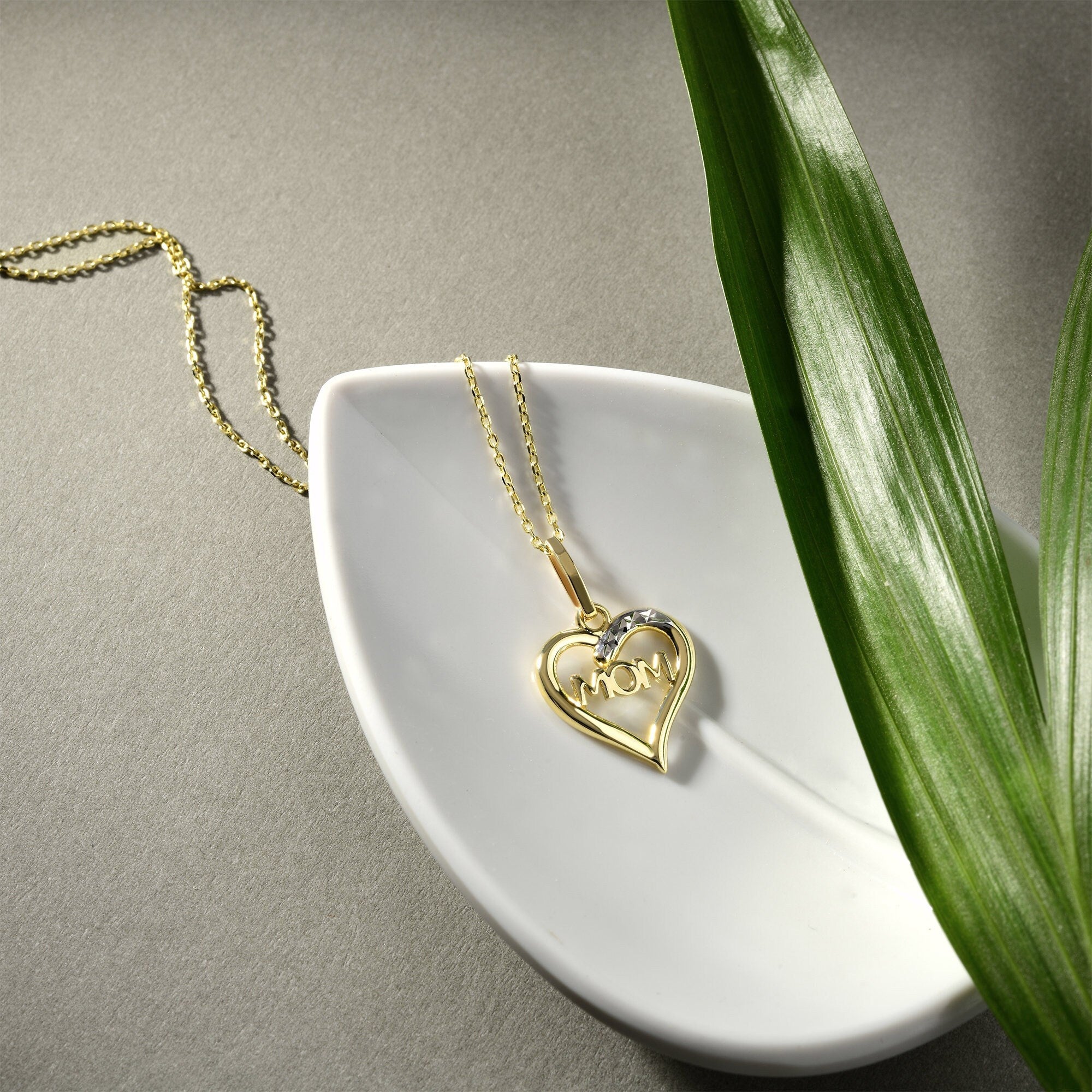 A 14K gold Mother Necklace from RundaJewelryUs, featuring a heart-shaped pendant with the word MOM, rests elegantly on a white, leaf-shaped dish. A green leaf beside the dish provides a striking contrast against the gray background.