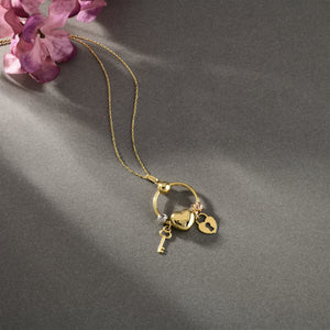 The 14K Solid Gold Heart Lock Key Necklace by RundaJewelryUs features a circular pendant adorned with three charms: a silver key, a gold heart, and a gold padlock with a keyhole. This pendant gracefully hangs from an elegant, delicate gold chain, capturing the timeless beauty of the lock and key design.