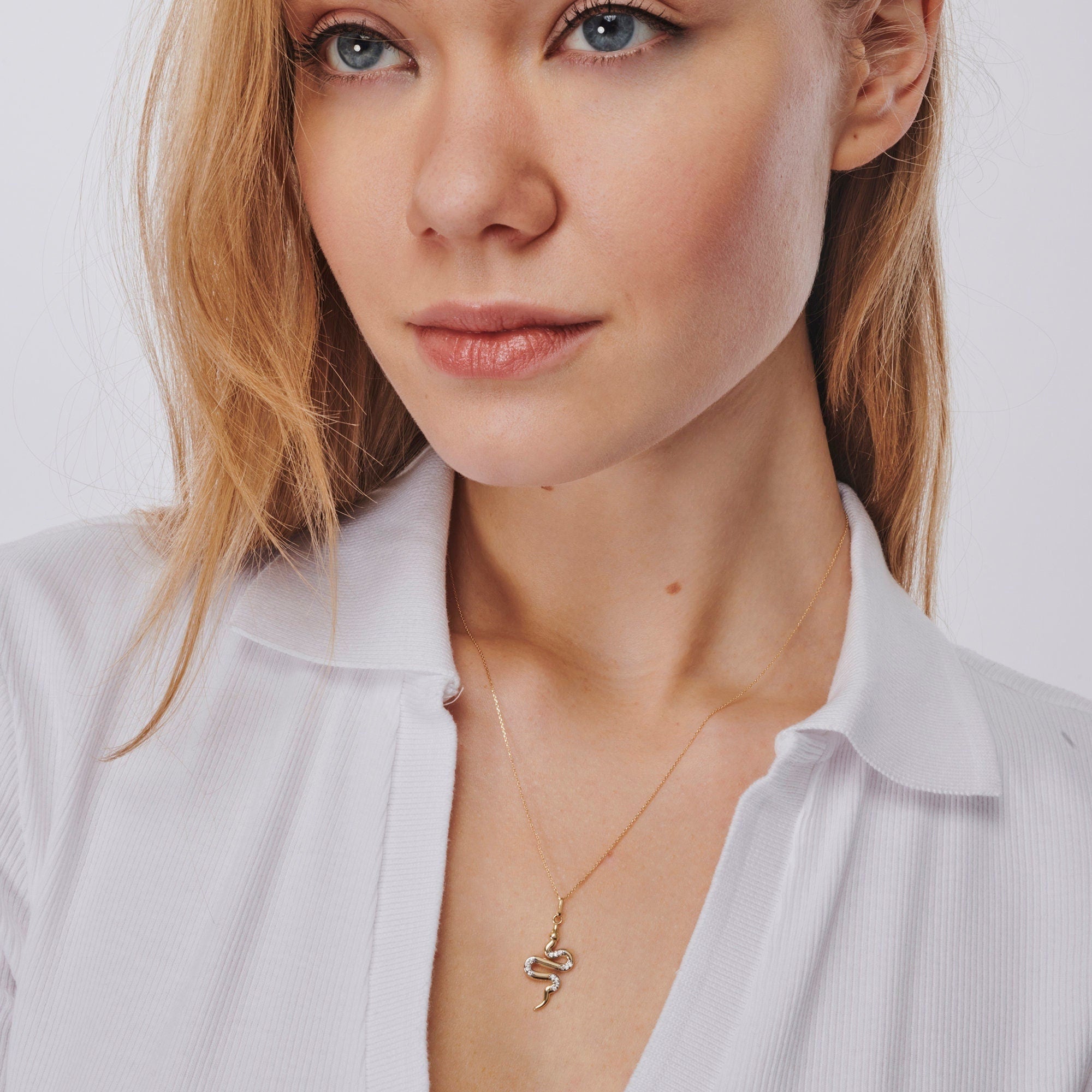 A person with long blonde hair is wearing a white blouse and a stunning RundaJewelryUs 14K Gold Snake Necklace for Women. The serpent necklace from RundaJewelryUs elegantly stands out, enhancing their neutral expression as they gaze slightly to the side.