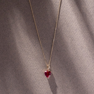 The 14K Gold Heart Shape Ruby Necklace showcases a heart-shaped red cubic zirconia gemstone pendant in a twisted gold setting. This exquisite piece features a fine chain with a simple link design and is crafted from solid 14K gold.