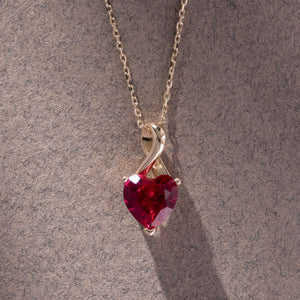 The 14K Gold Heart Shape Ruby Necklace showcases a heart-shaped red cubic zirconia gemstone pendant in a twisted gold setting. This exquisite piece features a fine chain with a simple link design and is crafted from solid 14K gold.