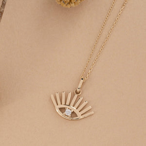 The 14K Solid Gold Evil Eye Necklace showcases a pendant in the shape of an eye, accented with sunburst rays extending above and below. At the center of this elegant design is a small round diamond, all suspended from a delicate gold chain. This piece serves as both a beautiful adornment and a symbol of protection.