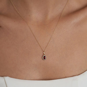 The 14K Gold Ruby Necklace showcases an exquisite oval-shaped red ruby encircled by a halo of sparkling diamonds. Its delicate chain enhances the pendant's refined elegance, while its hypoallergenic quality guarantees comfortable wear. The necklace is set against a plain, light background for added elegance.