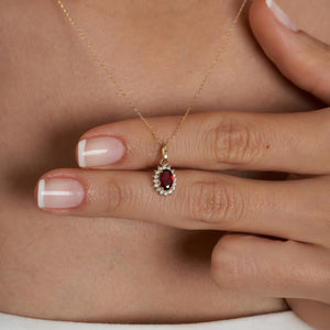 The 14K Gold Ruby Necklace showcases an exquisite oval-shaped red ruby encircled by a halo of sparkling diamonds. Its delicate chain enhances the pendant's refined elegance, while its hypoallergenic quality guarantees comfortable wear. The necklace is set against a plain, light background for added elegance.