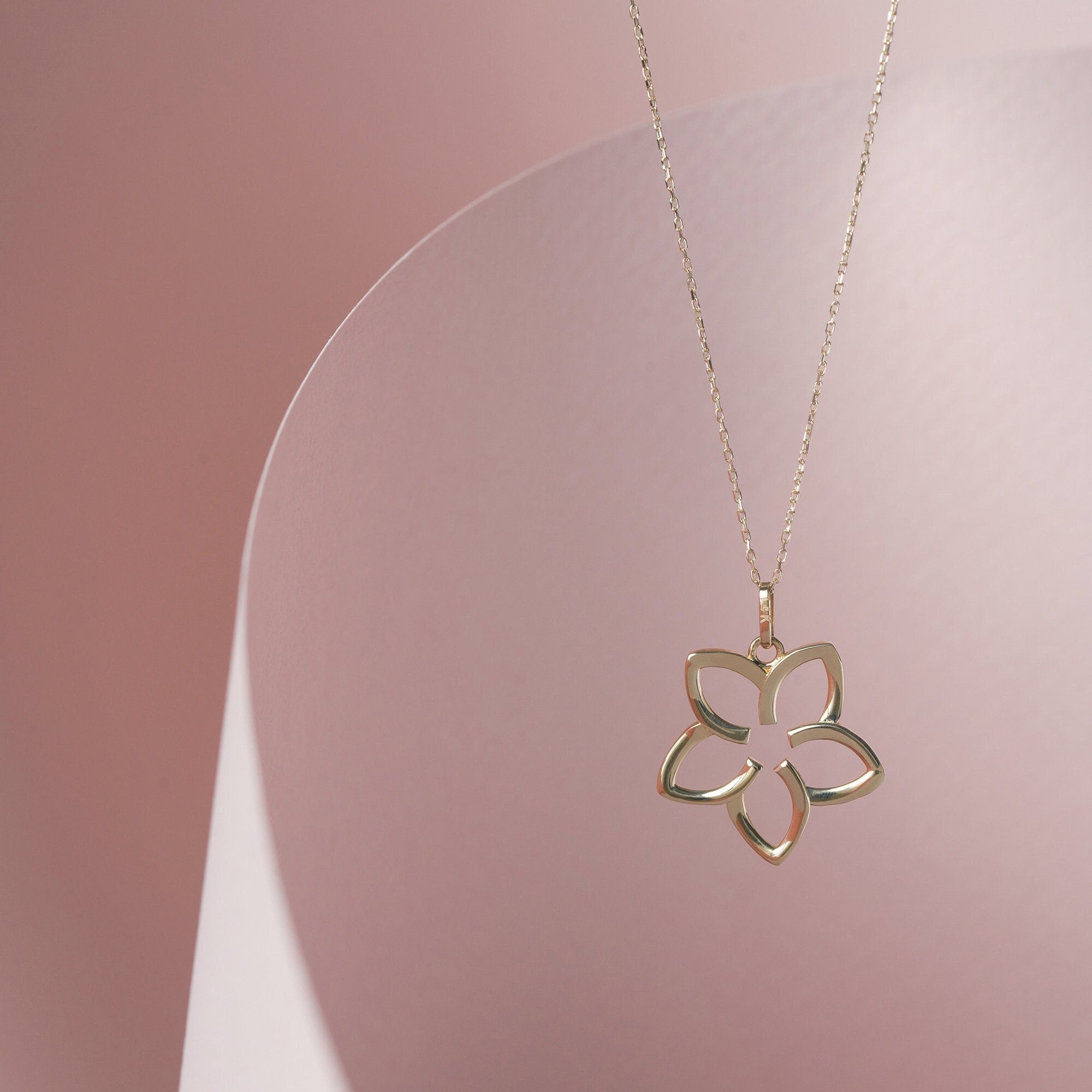 The 14K Gold Plumeria Necklace by RundaJewelryUs is gracefully showcased against a gentle pink, curved backdrop. The pendant's design highlights five open petals, representing new beginnings and offering a simple, stylish appearance.