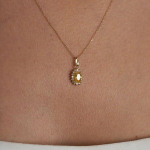 The 14K Gold Oval Citrine Necklace by RundaJewelryUs showcases a pendant with a prominent oval-shaped yellow citrine, encircled by a halo of shimmering white gemstones, elegantly displayed against a simple backdrop.