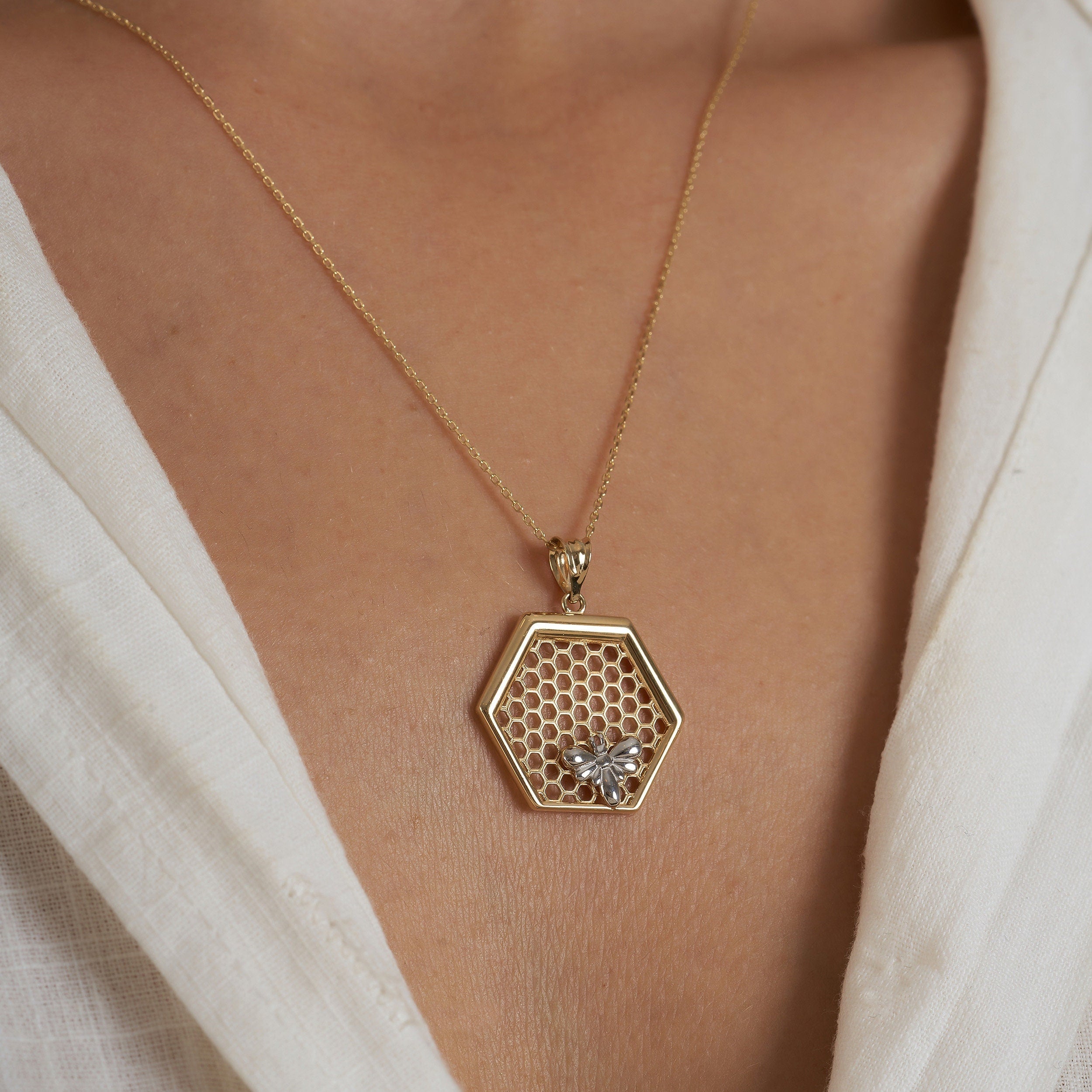 A person wearing the 14K Gold Queen Bee Pendant Necklace by RundaJewelryUs, featuring a hexagonal honeycomb pendant adorned with a delicate bee ornament. This exquisite piece of Honey Bee Jewelry is elegantly displayed on white textured fabric.