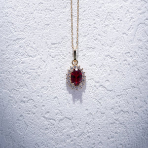 The 14K Gold Ruby Necklace showcases an exquisite oval-shaped red ruby encircled by a halo of sparkling diamonds. Its delicate chain enhances the pendant's refined elegance, while its hypoallergenic quality guarantees comfortable wear. The necklace is set against a plain, light background for added elegance.