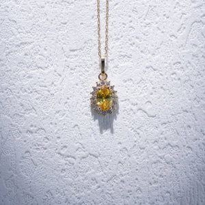 The 14K Gold Oval Citrine Necklace by RundaJewelryUs showcases a pendant with a prominent oval-shaped yellow citrine, encircled by a halo of shimmering white gemstones, elegantly displayed against a simple backdrop.