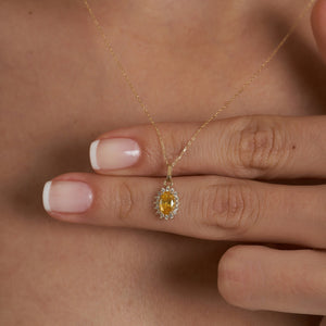 The 14K Gold Oval Citrine Necklace by RundaJewelryUs showcases a pendant with a prominent oval-shaped yellow citrine, encircled by a halo of shimmering white gemstones, elegantly displayed against a simple backdrop.