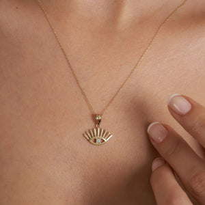 The 14K Solid Gold Evil Eye Necklace showcases a pendant in the shape of an eye, accented with sunburst rays extending above and below. At the center of this elegant design is a small round diamond, all suspended from a delicate gold chain. This piece serves as both a beautiful adornment and a symbol of protection.