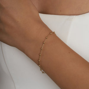 The 14K Solid Gold Bead Station Bracelet is a delicate piece, showcasing small round beads evenly spaced along its thin real solid gold chain. Set against a white background, this stunning bracelet is finished with a lobster clasp closure that ensures elegance and security.