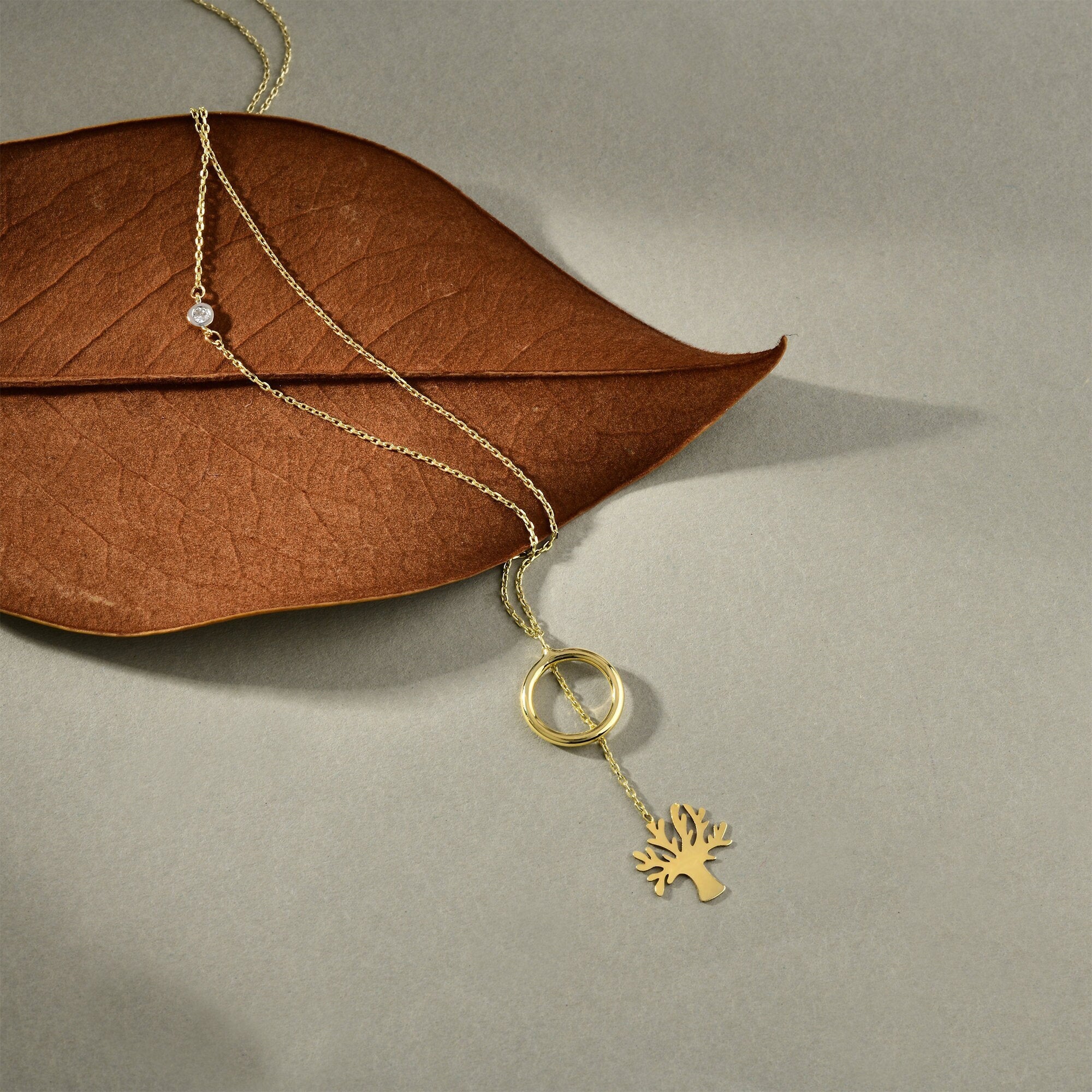 The 14k Solid Gold Tree of Life Y Necklace by RundaJewelryUs, featuring a tree-shaped pendant and a ring, is elegantly displayed on a brown leaf against a gray surface. Its simple, delicate design creates a gentle shadow.