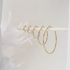 A pair of 14K Gold Round Hoop Earrings displayed on a plain white background features one earring shown from the side, showcasing its sleek, modern round shape and smooth finish, while the other is presented from the front, emphasizing its lightweight clasp mechanism.