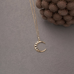 The 14K Real Gold Crescent Moon Phases Necklace showcases a crescent moon pendant embellished with subtle circular cutouts and shimmering cubic zirconia gemstones. Its delicate chain and the polished finish of the pendant contribute to its elegant appearance.