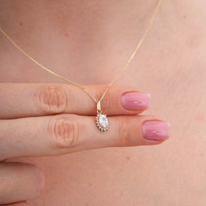 The 14K Gold Oval Cubic Zirconia Gemstone Necklace showcases a delicate design with an oval-shaped pendant adorned with a prominent cubic zirconia gemstone. Smaller stones encircle the centerpiece, creating a dazzling effect against a simple white backdrop.