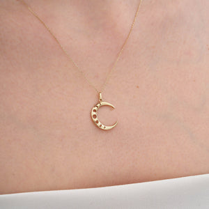 The 14K Real Gold Crescent Moon Phases Necklace showcases a crescent moon pendant embellished with subtle circular cutouts and shimmering cubic zirconia gemstones. Its delicate chain and the polished finish of the pendant contribute to its elegant appearance.