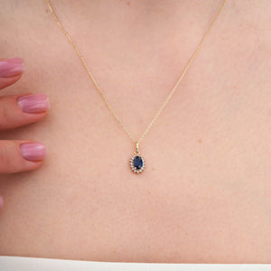 The 14K Gold Sapphire Necklace by RundaJewelryUs features a minimalist design, highlighted by an oval-shaped blue gemstone pendant surrounded by small white gemstones in a halo setting.