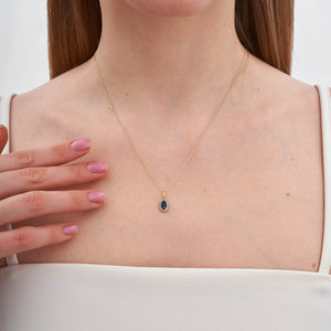 The 14K Gold Sapphire Necklace by RundaJewelryUs features a minimalist design, highlighted by an oval-shaped blue gemstone pendant surrounded by small white gemstones in a halo setting.