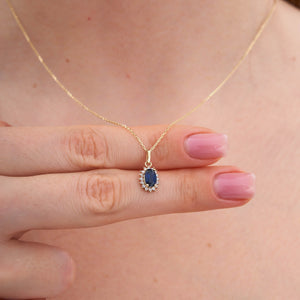 The 14K Gold Sapphire Necklace by RundaJewelryUs features a minimalist design, highlighted by an oval-shaped blue gemstone pendant surrounded by small white gemstones in a halo setting.