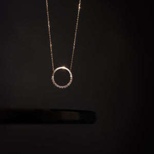 The 14K Gold Minimal Dainty Circle Necklace from RundaJewelryUs showcases a round pendant adorned with small diamonds embellishing the bottom half, producing a captivating sparkle. Set on a plain white background, its understated design accentuates the elegance and sophistication of this stunning jewelry piece.