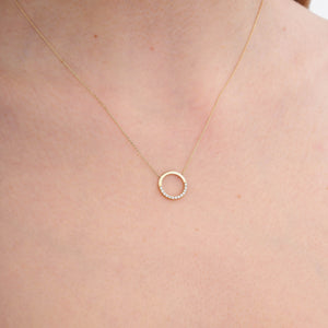 The 14K Gold Minimal Dainty Circle Necklace from RundaJewelryUs showcases a round pendant adorned with small diamonds embellishing the bottom half, producing a captivating sparkle. Set on a plain white background, its understated design accentuates the elegance and sophistication of this stunning jewelry piece.