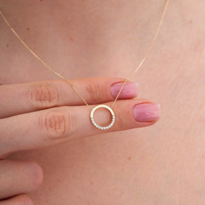 The 14K Gold Minimal Dainty Circle Necklace from RundaJewelryUs showcases a round pendant adorned with small diamonds embellishing the bottom half, producing a captivating sparkle. Set on a plain white background, its understated design accentuates the elegance and sophistication of this stunning jewelry piece.