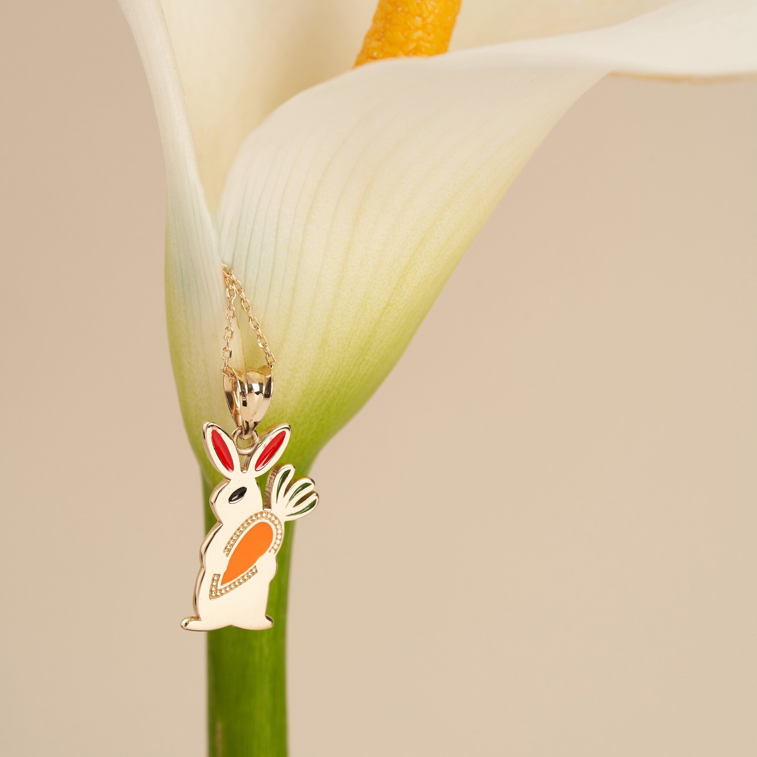 The 14K Gold Cute Rabbit Holding Carrot Necklace from RundaJewelryUs showcases a delicate pendant with a stylized bunny design in gold and orange. It hangs elegantly from a thin chain, beautifully displayed against the backdrop of a white calla lily on a neutral background.