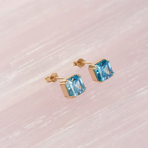 Presenting a stunning pair of 14K Gold Aquamarine Stud Earrings from Runda Jewelry. One earring is displayed from the front, emphasizing the vibrant aquamarine gemstone, while the other is shown from the side to beautifully showcase its elegant gold setting and post.