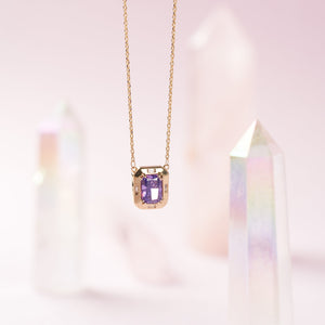 The 14K Solid Gold Amethyst Necklace, ideal for a February birthstone gift, showcases a rectangular pendant adorned with a vibrant purple gemstone. Surrounded by small, clear stones and hanging from a delicate chain, this handcrafted piece embodies elegance and charm.