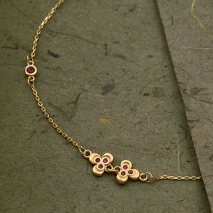 Introducing the 14K Solid Gold Ruby Leaf Bracelet from the Chloris Collection by RundaJewelryUs. This exquisite piece showcases a delicate chain design adorned with two small red floral motifs and three gold beads, reminiscent of the July birthstone. It features a secure lobster clasp and is elegantly finished with a circular tag at the end.