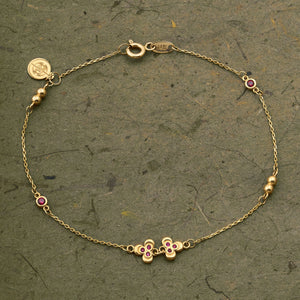 Introducing the 14K Solid Gold Ruby Leaf Bracelet from the Chloris Collection by RundaJewelryUs. This exquisite piece showcases a delicate chain design adorned with two small red floral motifs and three gold beads, reminiscent of the July birthstone. It features a secure lobster clasp and is elegantly finished with a circular tag at the end.