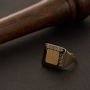 The 14K Gold Square Black Enamel Greek Key Signet Ring For Men features a square face embellished with a detailed black enamel Greek key pattern, echoing the labyrinthine meander design. Its bold, symmetrical style emphasizes the textured center, providing a stunning contrast between the black and gold elements.