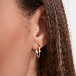 Displayed side by side are two elegant and minimalist hoop earrings from RundaJewelryUs. The left earring showcases a small diamond set in a flower-like design, while the right earring is part of the 14k Solid Gold Oval Solitaire Small Hoops collection, featuring a smooth, oval shape. Both earrings have a shiny finish that embodies the timeless allure of gold jewelry.