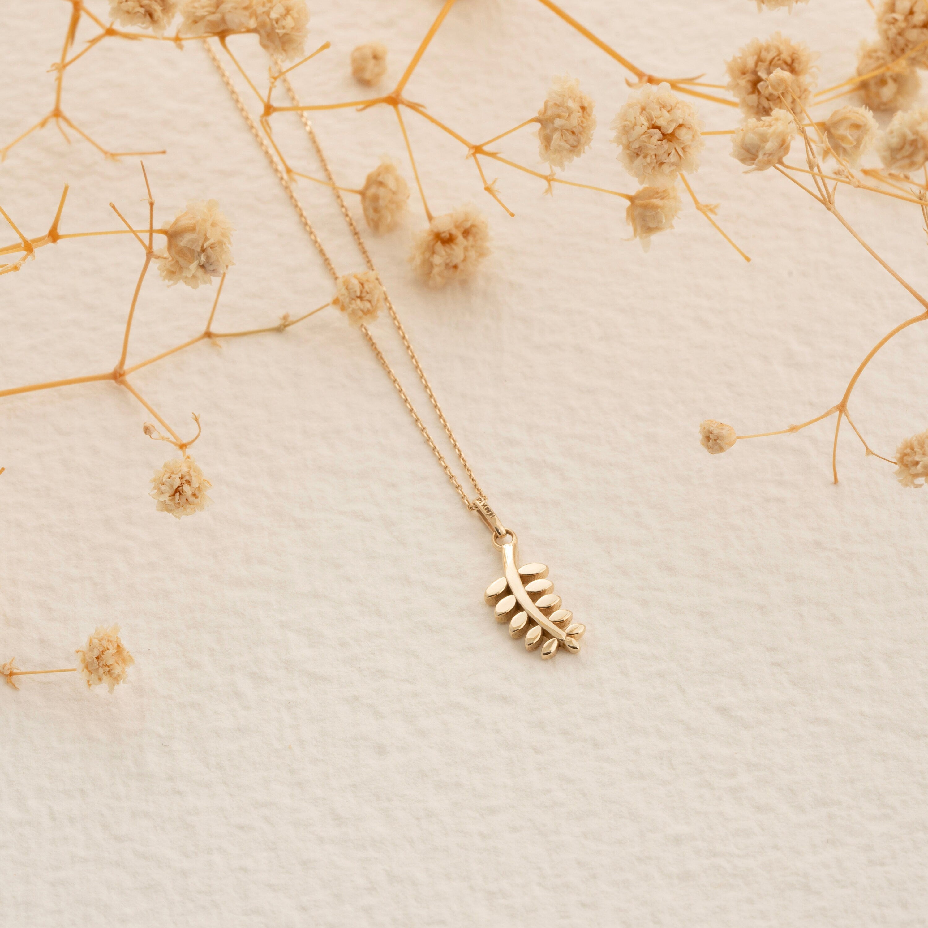 The 14K Solid Gold Dainty Leaf Necklace by RundaJewelryUs is a delicate handmade jewelry piece featuring a leaf-shaped pendant, beautifully set against a textured beige background. Its elegant and natural aesthetic is further enhanced by the artistic scattering of dried flowers around it.