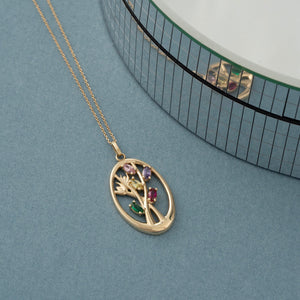 The 14K Solid Gold Birthstone Tree Pendant Necklace features a gorgeous pendant with an abstract tree design. It is set against a plain white background and showcases branches enhanced with multicolored gemstones in green, red, and purple, all enclosed within an oval frame.