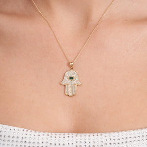 The 14K Solid Gold Emerald Hamsa Necklace showcases a pendant with a central eye-shaped green stone, providing protection against the evil eye. The design is beautifully intricate, highlighted by small clear stones, and complemented by a simple yet elegant chain.