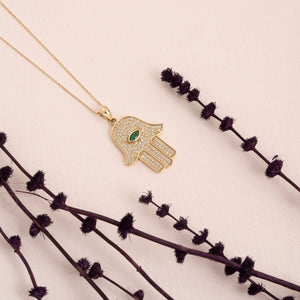 The 14K Solid Gold Emerald Hamsa Necklace showcases a pendant with a central eye-shaped green stone, providing protection against the evil eye. The design is beautifully intricate, highlighted by small clear stones, and complemented by a simple yet elegant chain.